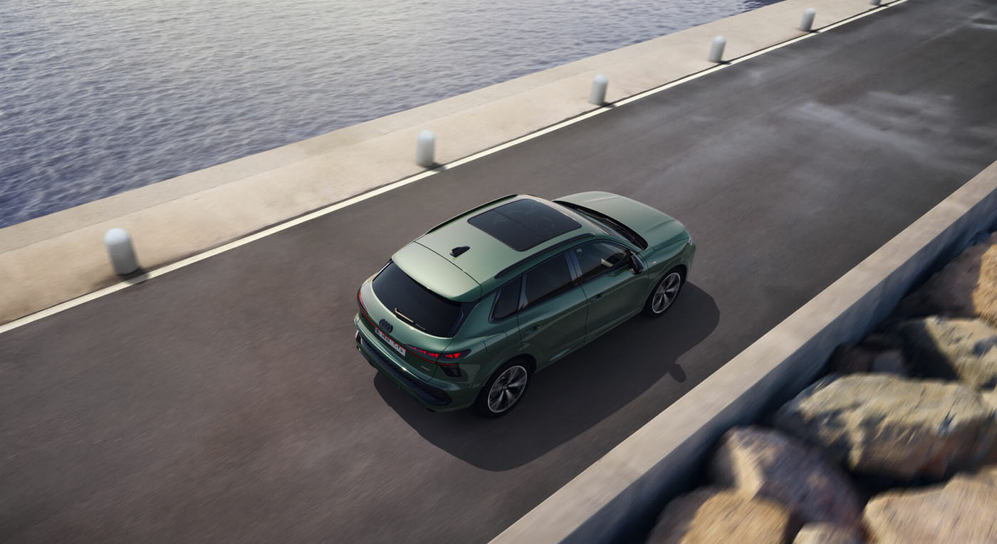 A green Audi Q3 SUV drives along a coastal road next to calm blue water. The setting conveys a sense of adventure and tranquility on a sunny day.